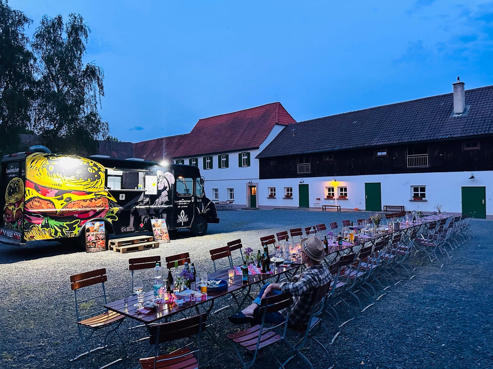 Lange gedeckte Tafel im Hof am Abend, daneben der beleuchtete Landstreicher-Truck
